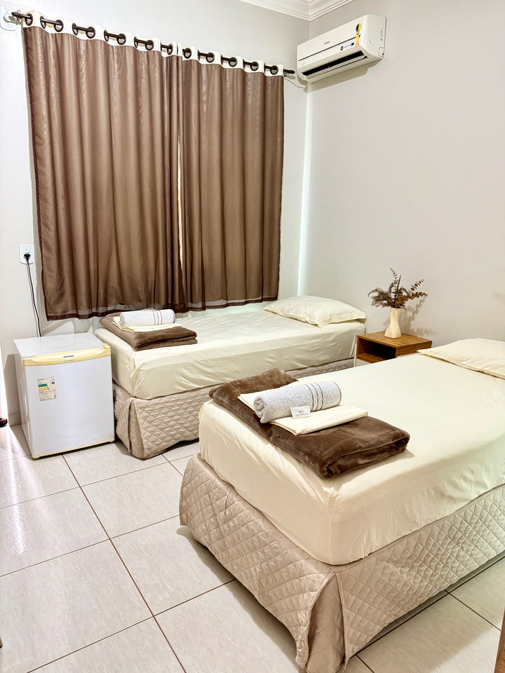 Duas camas de solteiro com toalhas dobradas, frigobar e cortina marrom